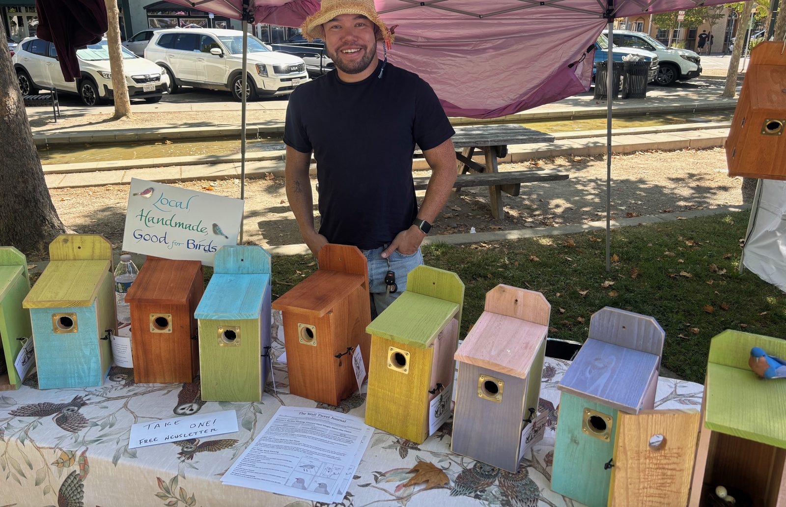 Adrian Baker-Kang behind the Bennett Valley Woodworks table at the farmers market, a row of painted cedar and redwood birdhouses in front of him.