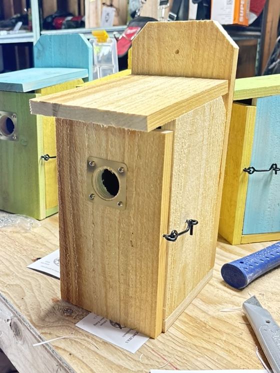 An unfinished birdhouse on the workbench showing the brass entry plate, hinged door and latch.